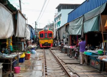 Colorful train moves through bustling Maeklong Railway Market in Bangkok, Thailand with local vendors.