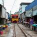 Colorful train moves through bustling Maeklong Railway Market in Bangkok, Thailand with local vendors.