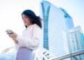 Asian woman using smartphone in Bangkok with modern skyscrapers in the background.